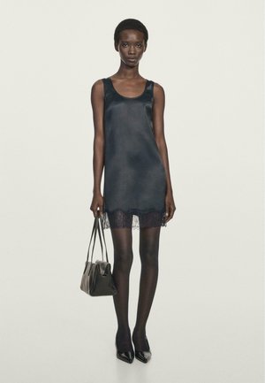 Black satin slip dress with lace hem, sleeveless design, worn with black sheer tights and pointed toe heels, held with a small black handbag.