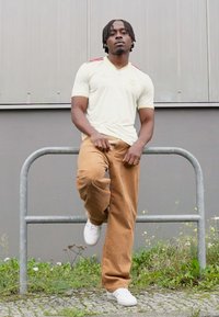 Maglietta sportiva bianca con dettagli rossi, pantaloni in corduroy marroni e sneakers bianche. Il modello si appoggia a un parapetto grigio, con uno sfondo neutro.