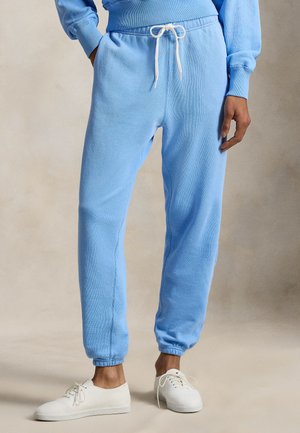 Persoană purtând pantaloni de trening albastru deschis cu șnur alb și adidași albi, stând în picioare pe un fundal neutru.