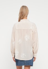 Camisa de manga longa bege claro com bordados florais brancos, punhos abotoados e barra curva, combinada com uma saia de denim. Vista de trás.