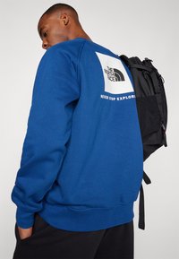 Sudadera azul con mangas largas y cuello redondo. Presenta un gran parche de logo en la parte posterior y una mochila negra colgada del hombro.