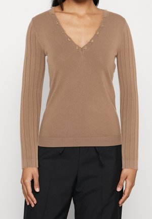 Kvinde iført en tætsiddende kamelfarvet V-hals sweater med dekorative løkker og ribstrikkede lange ærmer, kombineret med sorte bukser.