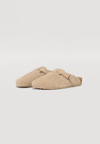 Chaussons en daim beige avec un bout arrondi, un dos ouvert et une boucle de sangle sur le côté. Matériau texturé et semelle en caoutchouc pour une bonne adhérence.