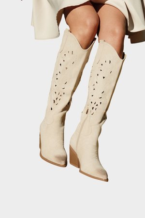 Bottes en daim beige montant jusqu'aux genoux avec des motifs découpés au laser, bouts pointus et talons superposés, portées avec une jupe légère.