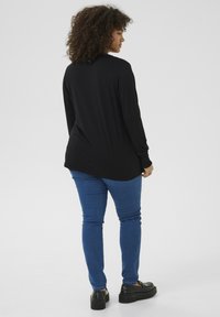 Haut noir à manches longues avec un ourlet incurvé, associé à un jean slim bleu et des chaussures à plateforme noires, vu de l'arrière.