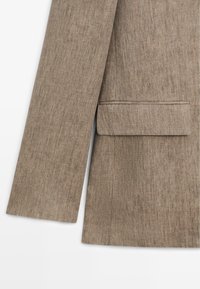 Mouw en onderste helft van een bruine getextureerde blazer met een klepzak op een witte achtergrond.