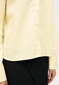 Gros plan d'une personne portant une chemise jaune pâle à boutons avec manches longues et une jupe noire avec un panneau avant chevauchant.