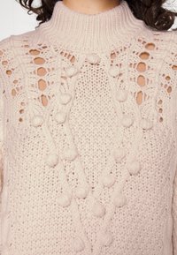 Maglione intrecciato rosa chiaro con colletto alto, caratterizzato da motivi a doppio gioco e dettagli testurizzati in rilievo.