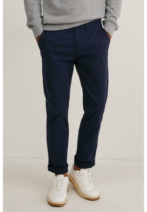 Man draagt marineblauwe opgerolde broek, grijze trui en witte sneakers, staand met handen in de zakken.