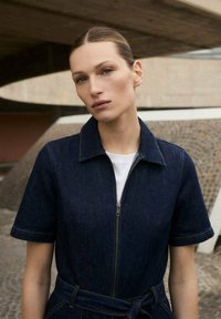 Mono de mezclilla oscuro con manga corta, cuello, cierre frontal de cremallera y cintura ajustada con tiras. Lleva una camisa blanca debajo, con una textura suave.