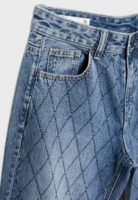 Ljusblå denimjeans med ett strukturerat diamantmönster skapat av små metalliska nitar; har en framficka och standard midjeband.