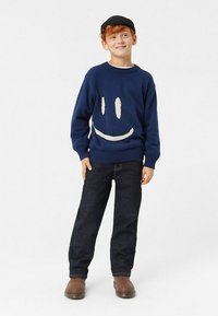 Tummansininen neulehuppu, jossa on valkoinen hymyilevä kasvomuotoilu, yhdistettynä tummiin farkkuihin ja ruskeisiin saappaisiin. Yksinkertainen, rento asu.