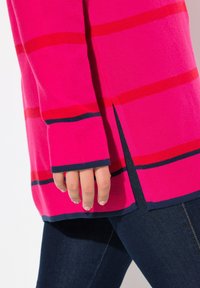 Sweter w kolorze fuksji w pionowe paski w czerwonym i granatowym kolorze, z prążkowaną teksturą i rozcięciami po bokach. Zestawiony z ciemnymi dżinsami.