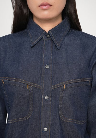 Tmavomodrá denimová košeľa so zapínaním na gombíky, špicatým golierom a dvoma náprsnými vreckami. Má viditeľné švy v kontrastnej žltej farbe.