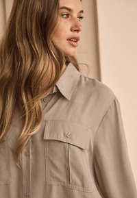Light beige button-up shirt with a collar and two flap pockets. Smooth fabric with subtle sheen, featuring stitch detailing along seams.