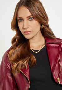 Giubbotto in pelle bordeaux con zip sul davanti, indossato sopra un top nero a coste. Una collana in tonalità oro e orecchini a cerchio completano il look.