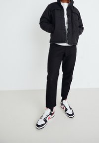Person som bär en svart pufferjacka, svarta byxor, vit skjorta och svarta, vita och röda Nike-sneakers som står mot en enfärgad bakgrund.