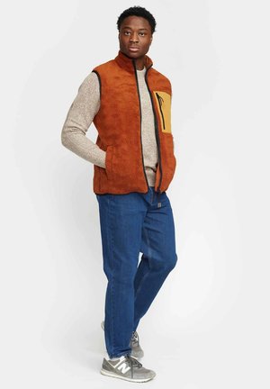 Uomo con gilet in pile arancione con tasca gialla sul petto, maglione beige, jeans blu e sneakers grigie, in piedi con le mani nelle tasche.