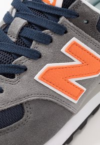 Tenis gri din suede, cu plasă albastră închisă, accent logo portocaliu, detalii albe și talpă din cauciuc negru cu textură.