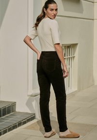 Šviesiai smėlinės spalvos raukšlėta trumpa rankovė, juodos aukštu juosmeniu džinsai ir rudos spalvos šlepetės. Modelis pozuoja su ranka ant klubo, plaukai surišti.