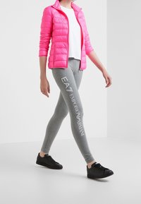 Rosa puffjacka, vit t-shirt, grå leggings med tryckt logotyp och svarta sneakers. Materialen är blanka, stretchiga och släta.