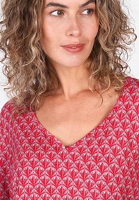 V-Ausschnitt-Oberteil aus weichem Stoff, mit rotem Grundton und aufwendigen floralen Mustern in Rosa und Grün. Lockige Haare sind am Ausschnitt sichtbar.