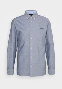 Valkoinen painatettu logolla varustettu vaaleansininen kauluspaita, joka on valmistettu chambray-kankaasta. Paidassa on pitkät hihat, terävä kaulus ja valkoiset napit.