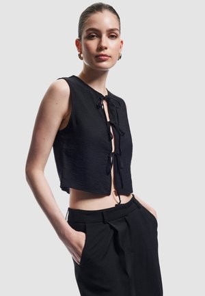 Schwarzes Cropped Top mit strukturierter Oberfläche und vorderem Bindedetail, kombiniert mit hoch sitzenden schwarzen Hosenträgern mit Seitentaschen.