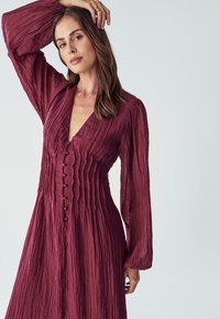 Burgundy, texturované šaty s dlouhým rukávem, V výstřihem a detaily knoflíčků, s volánky probíhajícími svisle od výstřihu až po lem.