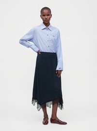 Femme portant une chemise rayée bleue à boutons et une jupe asymétrique noire ornée de dentelle, avec des chaussures plates bordeaux.