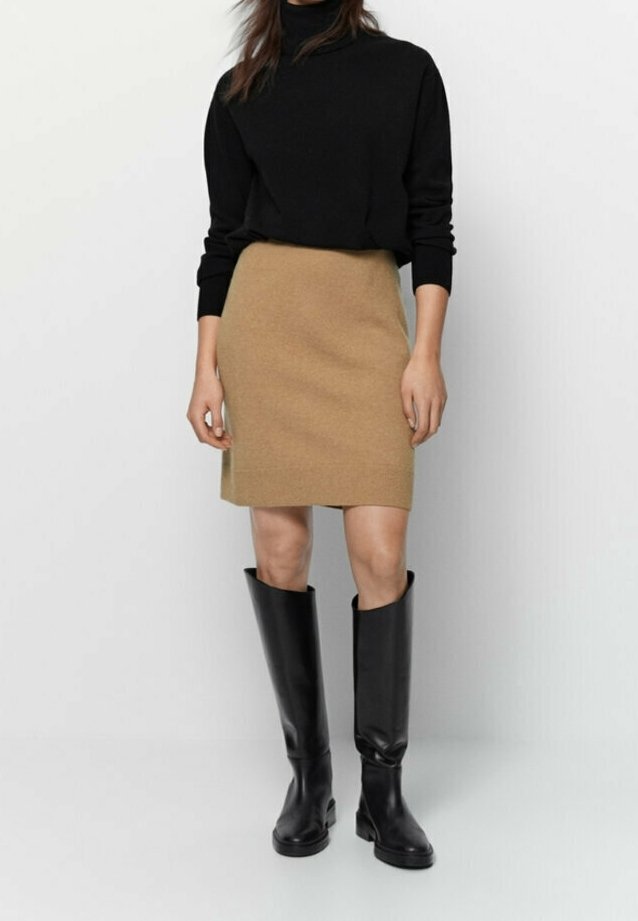Femme portant un pull à col roulé noir, une jupe couleur camel longueur genou et des bottes en cuir noires hauteur genou, debout devant un fond uni.
