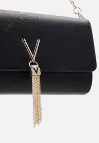 Svart läderclutch med texturerad yta, som har en guldmetall-emblem och kedja med tassel-detalj för extra dekoration.