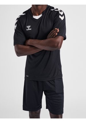 Mann steht mit verschränkten Armen, trägt ein schwarzes hummel Sportshirt und Shorts vor einfachem grauem Hintergrund.