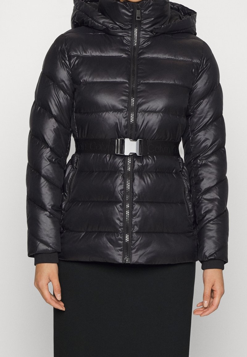 Femme portant une doudoune noire matelassée avec capuche, fermeture éclair frontale, poches zippées et boucle de ceinture de marque à la taille.