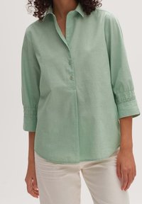 Ljusgrön skjorta med knappar och krage, trekvartslånga ärmar med elastiska manschetter och en böjd fåll. Tillverkad av ett mjukt, strukturerat tyg.