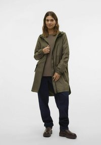 Giacca impermeabile verde oliva con cappuccio, polsini elastici, tasche frontali e zip. Indossata sopra un maglione marrone e jeans blu larghi.