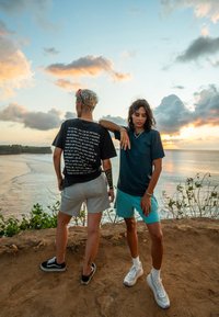 Dvoje pojedinaca stoji na litici. Jedan nosi crnu majicu s grafikom i sive šorts, dok drugi nosi tamnu majicu i svijetloplave šorts.