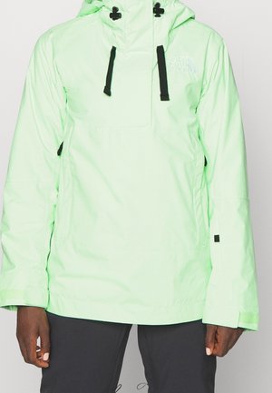 Chaqueta ligera verde con capucha para exteriores con cordones negros, logo de The North Face en el pecho, usada con pantalones negros.
