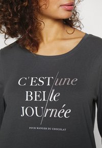 Dark gray, long-sleeve shirt featuring a round neckline and printed text in white and silver: "C'EST une BELLE JOURnée." Smooth texture.
