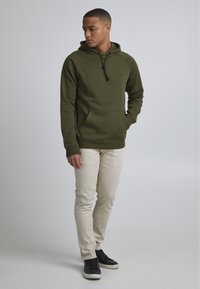 Sudadera verde oliva con un bolsillo tipo canguro, capucha con cordón y puños acanalados, combinada con pantalones beige claros y zapatillas negras.