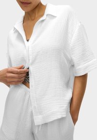 Camicia bianca a maniche corte con bottoni, realizzata in tessuto testurizzato, caratterizzata da un taglio rilassato e un colletto. Abbinata a pantaloni bianchi coordinati.