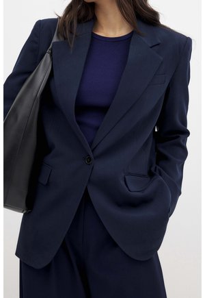 Woman wearing a navy blazer and matching pants over a ribbed navy top, carrying a large black leather shoulder bag, hand in pocket.