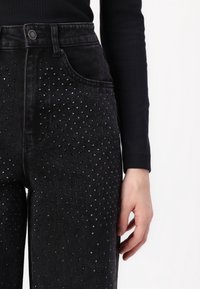 Jean en denim noir au design ajusté, avec des clous argentés décoratifs disposés en motif de points sur le devant.