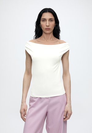 Femme aux longs cheveux noirs portant un haut blanc épaules dénudées et un pantalon violet clair, se tenant devant un fond clair uni.