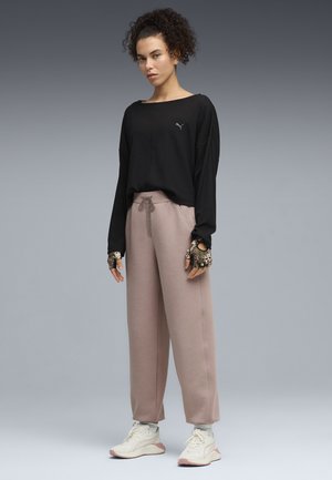 Jong persoon staand in casual sportkleding: zwart langermig topje, roze broek met trekkoord, witte sneakers en vingerloze handschoenen.