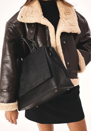 Personne portant une veste en peau retournée marron foncé et une jupe noire, tenant un grand sac à main noir texturé avec des pieds en métal et des détails en chaîne.