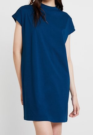 Femme portant une robe bleue unie à manches courtes, longueur genoux, avec un col rond haut, debout devant un fond clair.