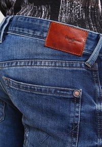 Modré džíny z denimu s hnědou koženou nášivkou označenou "Pepe Jeans." Mezi zvláštnosti patří kontrastní švy a kovové detaily knoflíků.