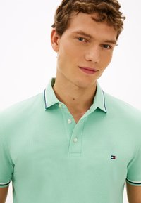 Ανοιχτό πράσινο πόλο μπλουζάκι με υφή υφάσματος, διαθέτει δύο κουμπιά και λεπτομέρειες σε ναυτικό μπλε στο γιακά και τα μανίκια. Κεντημένο λογότυπο.