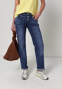 Personne portant un jean bleu retroussé, un haut jaune sans manches, des baskets argentées, tenant un grand sac marron souple dans la main gauche.